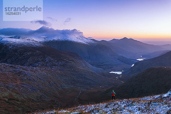 Mann in orange-blauer Jacke steht im Morgengrauen des Winters auf einem schneebedeckten Hügel mit Blick auf das Black Valley und die Carrauntoohil; Grafschaft Kerry  Irland