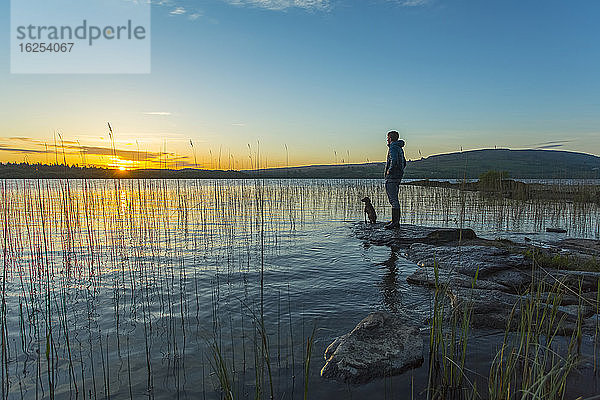 Mann und Hund stehen auf einem Felsen am Rande des Lough Graney und beobachten den Sonnenaufgang; Grafschaft Clare  Irland