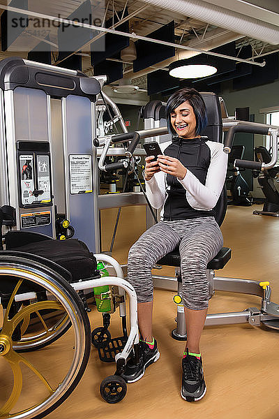 Eine querschnittsgelähmte Frau  die sich nach dem Training mit einer Überkopfpresse in einer Fitnesseinrichtung ausruht und mit ihrem Smartphone SMS schreibt; Sherwood Park  Alberta  Kanada