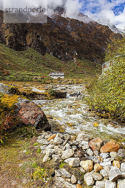 Ein Fluss fließt an einem Teehaus mit Gebetsfahnen vorbei  an einem Felsblock mit orangefarbenen Gebeten  die auf den Felsen geschrieben sind  an einem sonnigen Herbsttag mit frischem Schnee auf den Gipfeln im Hintergrund  während sich die Wolken auflösen  außerhalb des Dorfes Dole  Sagarmatha-Nationalpark; Dole  Nepal