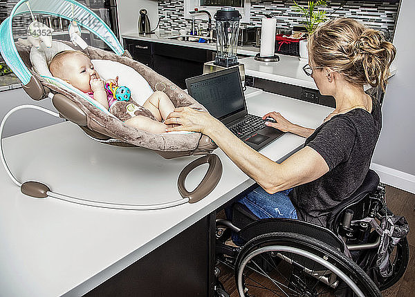 Eine junge Mutter mit einer Rückenmarkverletzung kümmert sich um ihr Neugeborenes  während sie in der Küche an ihrem Computer arbeitet; Edmonton  Alberta  Kanada