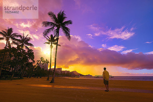 Ein Mann steht am Strand von Waikiki und blickt bei Sonnenaufgang auf die Eigentumswohnungen und Palmen entlang der Küstenlinie von Waikiki; Honolulu  Oahu  Hawaii  Vereinigte Staaten von Amerika