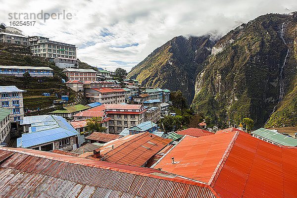 Blick den Berg hinunter auf das Dorf Namche Bazaar  ein wichtiger Zwischenstopp und oft als Zwischenstopp genutzt  entlang des Everest- und Gokyo-Treks  zu all den farbenfrohen Teehäusern  an einem bewölkten Herbstmorgen  Sonnenlicht küsst die Berge auf der anderen Seite des Tals; Namche Bazaar  Nepal