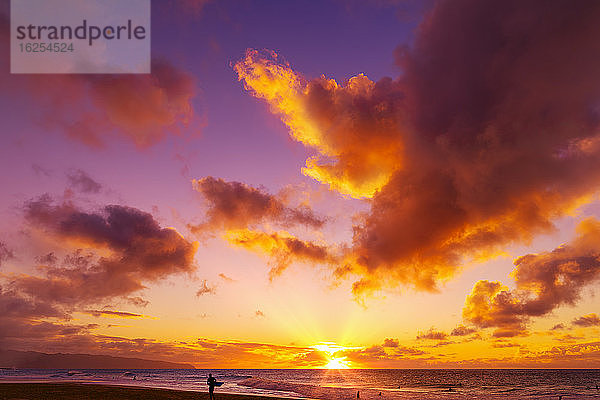 Silhouette eines Surfers  der am Kelki Beach am Wasser steht  mit dramatisch leuchtenden Wolken darüber bei Sonnenuntergang; Oahu  Hawaii  Vereinigte Staaten von Amerika