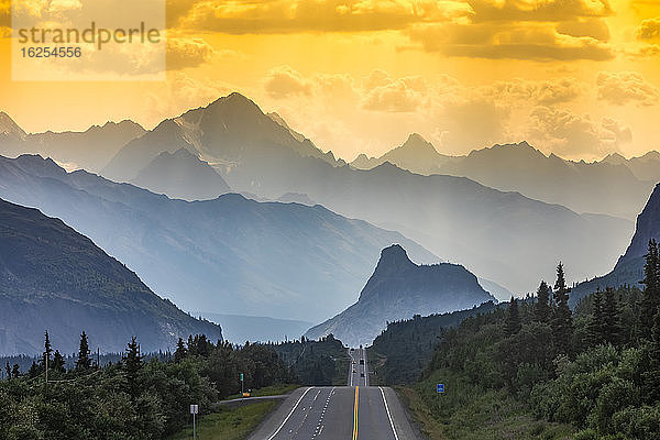 Glenn Highway teilt das Bild  umgeben von Fichtenwald und Gestrüpp im südlichen Zentralalaska  bei Sonnenuntergang  mit Autos in der Ferne  die in einer verrauchten  dunstigen Sommernacht auf Lion's Head und die Chugach-Berge blicken; Chickaloon  Alaska  Vereinigte Staaten von Amerika