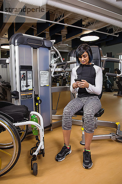 Eine querschnittsgelähmte Frau  die sich nach dem Training mit einer Überkopfpresse in einer Fitnesseinrichtung ausruht und mit ihrem Smartphone SMS schreibt; Sherwood Park  Alberta  Kanada