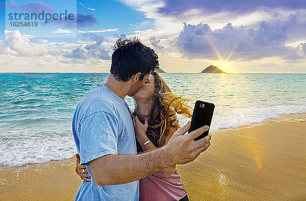 Ein Paar steht küssend am Strand von Lanakai auf der hawaiianischen Insel Oahu  während es ein Smartphone in der Hand hält und bei Sonnenuntergang ein Selbstporträt aufnimmt; Lanakai  Oahu  Hawaii  Vereinigte Staaten von Amerika