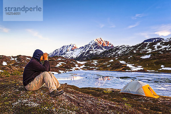 Ein Mann  der bei Sonnenuntergang mit einem Fernglas aus dem Lager am Lost Lake schaut. Gipfel der Auferstehung im Hintergrund. Chugach National Forest  Kenai-Halbinsel  Süd-Zentral-Alaska im Frühling; Seward  Alaska  Vereinigte Staaten von Amerika