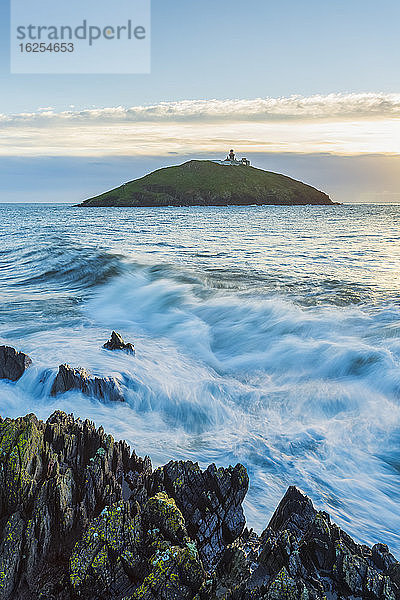 Wellen prallen bei Sonnenaufgang gegen die Küstenfelsen mit dem Leuchtturm Ballycotton im Hintergrund; Ballycotton  Grafschaft Cork  Irland