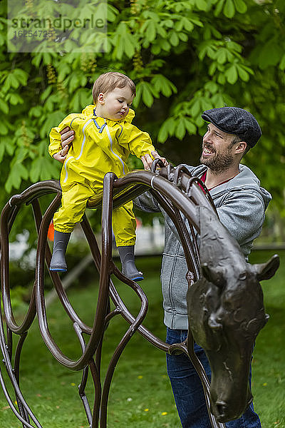 Nahaufnahme eines Vaters  der seine Tochter auf einer Pferdeskulptur im Park an einem bedeckten Frühlingstag festhält; North Vancouver  British Columbia  Kanada