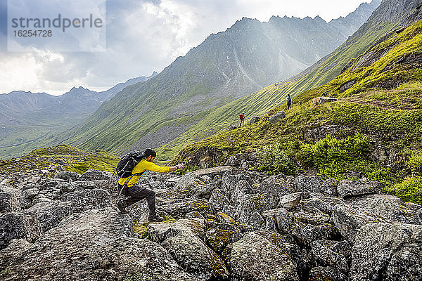 Ein kaukasischer Mann springt von Felsbrocken zu Felsbrocken über ein Geröllfeld  mit einem asiatischen Mann und einer kaukasischen Frau und ihrer Hundewanderung im Hintergrund auf dem sehr felsigen/geröllreichen Reed Lakes-Trail durch die Talkeetna-Berge am Hatcher's Pass  Palmer  Alaska  am späten Nachmittag im Sommer; Palmer  Alaska  Vereinigte Staaten von Amerika