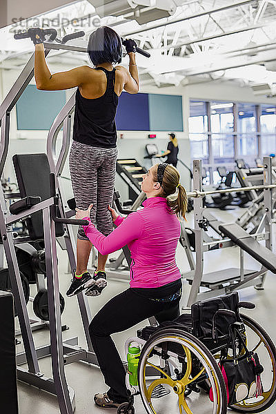 Eine Physiotherapeutin unterstützt eine querschnittsgelähmte Frau  die Klimmzüge macht  um ihre Oberkörperkraft zu erhöhen  in einer Fitnesseinrichtung: Sherwood Park  Alberta  Kanada