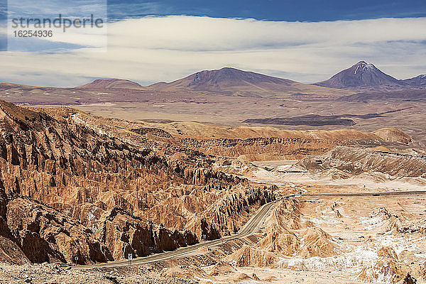 Die Straße führt hinunter in ein hochgelegenes Wüstental mit einzigartigen Felsformationen auf der linken Seite und einem Vulkangipfel in der Ferne; San Pedro de Atacama  Antofagasta  Chile