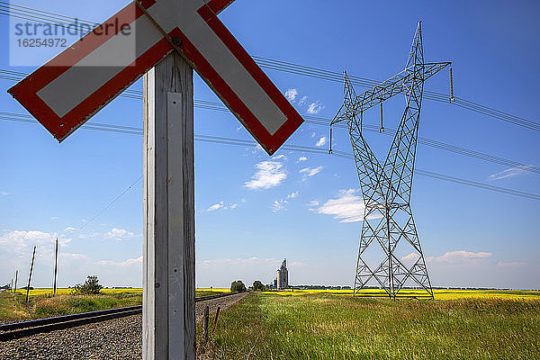 Nahaufnahme eines Bahnübergangsschildes und eines Sendeturms vom Typ Taille in einem Rapsfeld mit Zuggleisen und Getreideterminal in der Ferne; Alberta  Kanada