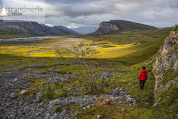 Eine Frau in rotem Mantel geht auf das Sonnenlicht zu  das die Tundra nach einem Regensturm im Spätsommer erhellt  Upper Marsh Fork Valley  Brooks Range  Arctic National Wildlife Refuge; Alaska  Vereinigte Staaten von Amerika