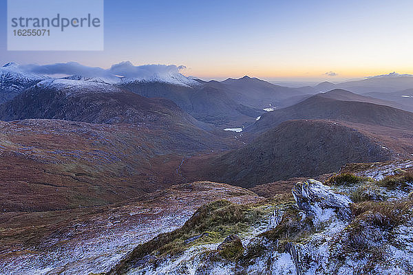 Felsvorsprünge und Heidekraut  die an einem Berghang hinunter zu den Seen des Black Valley führen  mit den MacGillycuddy's Reeks im Hintergrund in der Morgendämmerung im Winter; Grafschaft Kerry  Irland