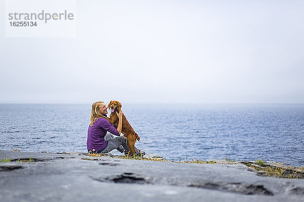 Frau umarmt Hund auf felsigem Grund mit Blick auf das Meer an einem bewölkten Tag; Fanore  Grafschaft Clare  Irland