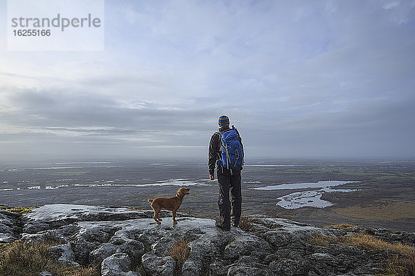 Mann mit Rucksack und Hund steht auf einer Klippe mit Blick auf die Seen im Burren-Nationalpark; Grafschaft Clare  Irland