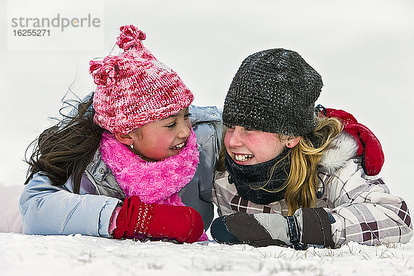 Zwei Mädchen in Winterkleidung liegen auf dem schneebedeckten Boden und lächeln sich an; Alberta  Kanada