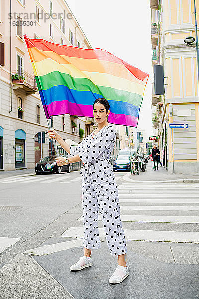 Junge lesbische Frau steht auf einer Straße und schwenkt eine Regenbogenfahne.