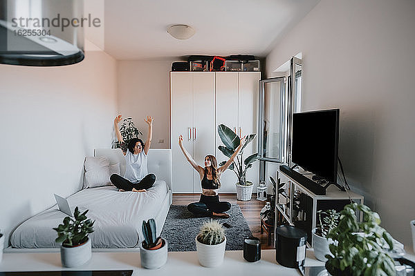 Zwei Frauen mit braunem Haar sitzen in einer Wohnung und machen Yoga.