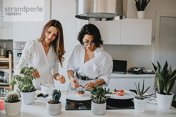 Zwei lächelnde Frauen mit braunem Haar stehen in einer Küche und bereiten Essen zu.
