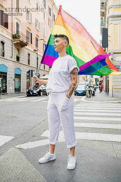 Junge lesbische Frau steht auf einer Straße und schwenkt eine Regenbogenfahne.
