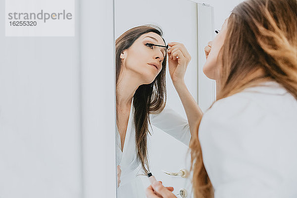 Frau mit braunem Haar steht vor einem Spiegel und trägt Wimperntusche auf.