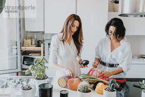 Zwei lächelnde Frauen mit braunem Haar stehen in einer Küche und nehmen Gemüse aus dem Einkaufsnetz.
