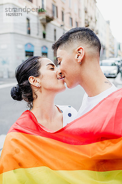 Porträt eines jungen lesbischen Paares  das auf einer Straße steht  in eine Regenbogenfahne gehüllt  und sich küsst.