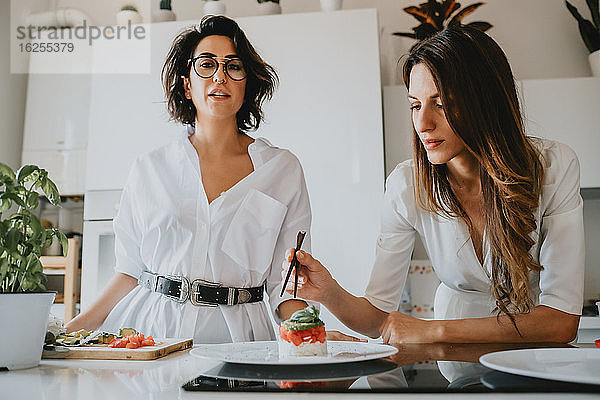 Zwei lächelnde Frauen mit braunem Haar stehen in einer Küche und bereiten Essen zu.