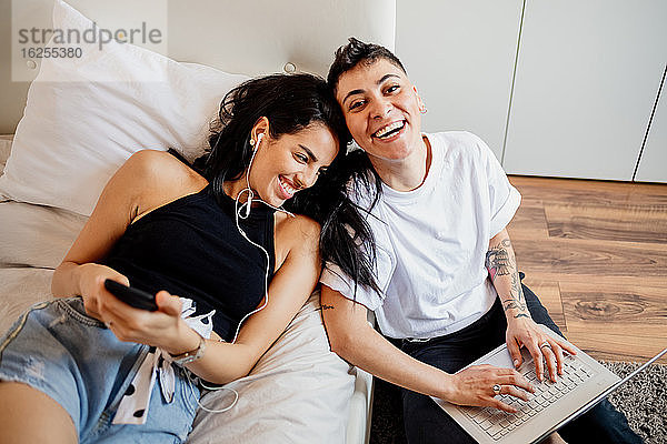 Junges lesbisches Paar in einem Schlafzimmer  mit Handy und Laptop in der Hand  lächelt in die Kamera.