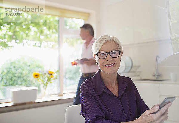 Porträt einer glücklichen älteren Frau  die ein Smartphone in einer sonnigen Küche benutzt