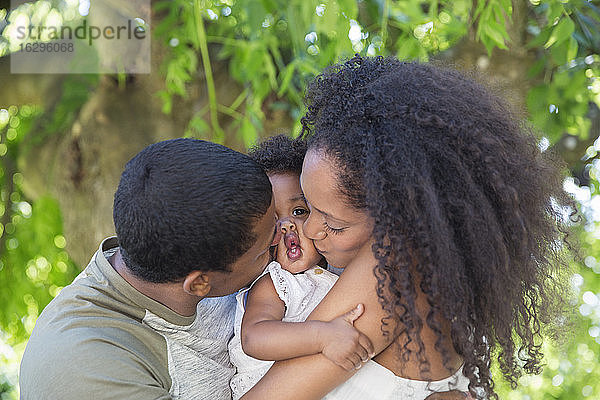 Liebevolle Eltern küssen Kleinkind-Tochter unter Baum