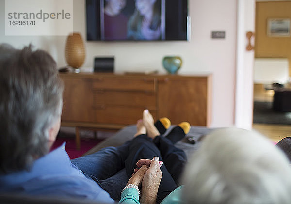 Älteres Ehepaar hält Händchen und schaut auf dem Wohnzimmersofa fern