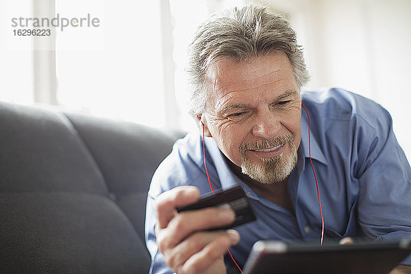 Älterer Mann mit Kopfhörer und Kreditkarte mit digitalem Tablett auf dem Sofa