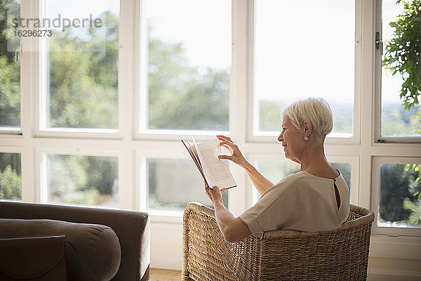 Ältere Frau entspannt sich und liest ein Buch im sonnigen Wohnzimmer