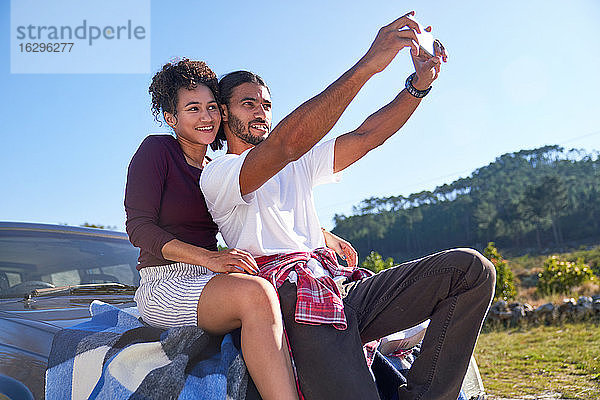 Glückliches junges Paar beim Selbstfahren auf der Motorhaube am sonnigen Straßenrand