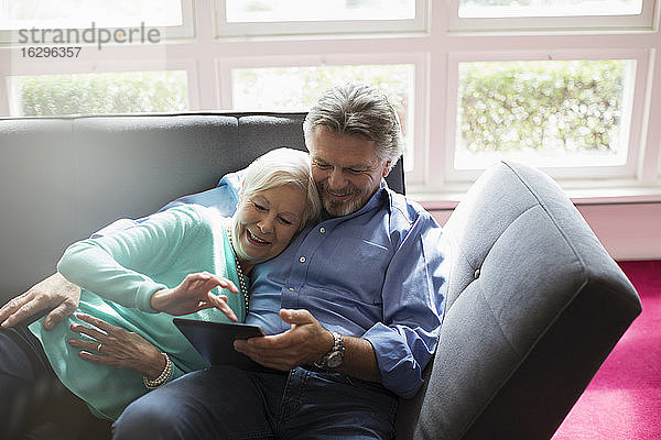 Glückliches Seniorenpaar kuschelt und benutzt digitales Tablet auf dem Sofa