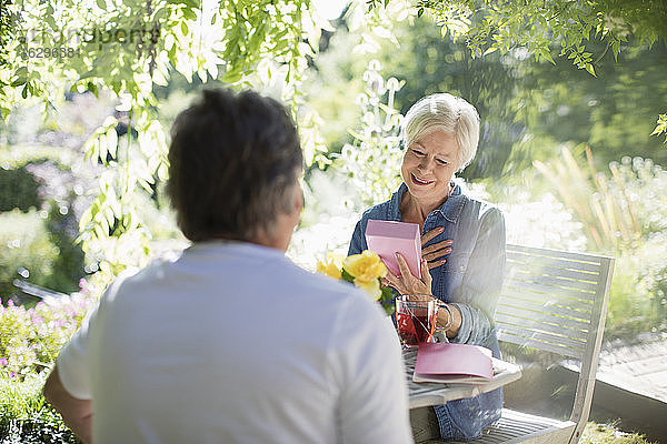 Glückliche ältere Frau öffnet Geschenk von Ehemann auf sonniger Sommerterrasse