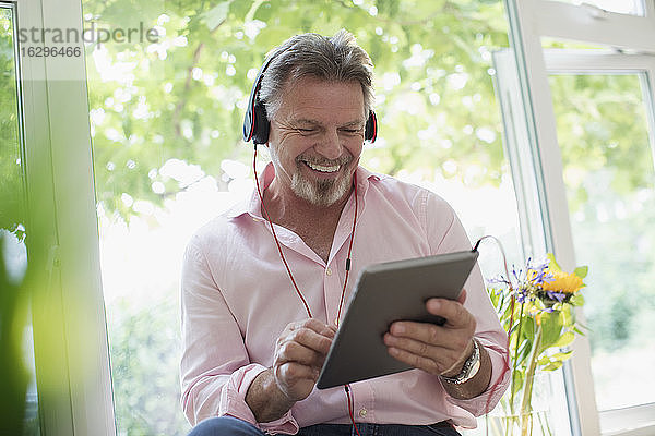 Glücklicher älterer Mann mit Kopfhörer und digitalem Tablet am Fenster