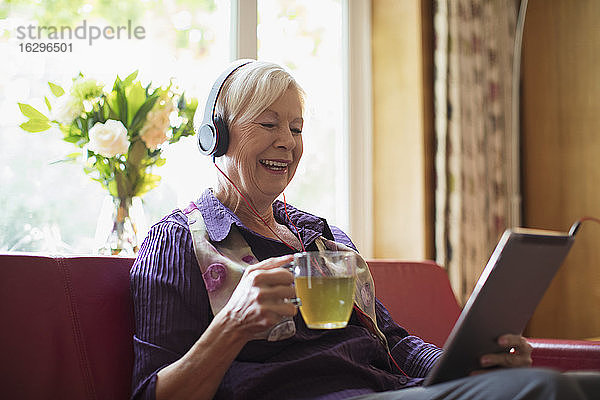 Glückliche ältere Frau mit Kopfhörern und digitaler Tablette beim Teetrinken