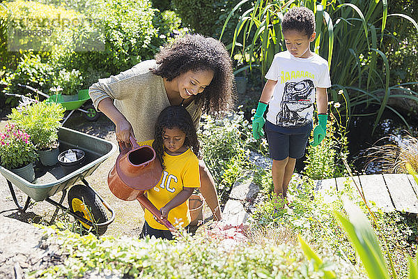 Glückliche Mutter und Kinder gießen Pflanzen im sonnigen Sommergarten