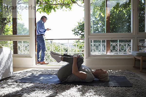 Ältere Frau  die sich auf einer Yogamatte vor der Balkontür im Sommer streckt