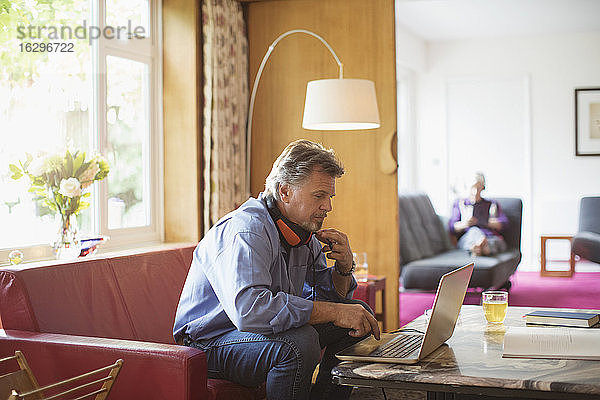 Älterer Mann mit Kopfhörern arbeitet am Laptop auf dem Wohnzimmersofa