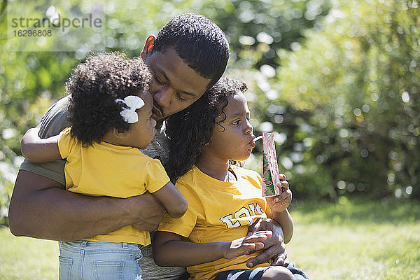 Liebevoller Vater umarmt Töchter im sonnigen Sommerhinterhof