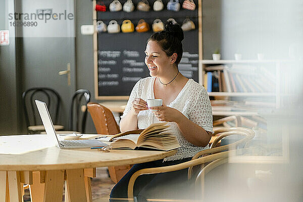 Lächelnde junge Frau hält eine Kaffeetasse und schaut auf einen Laptop auf dem Tisch in einem Café