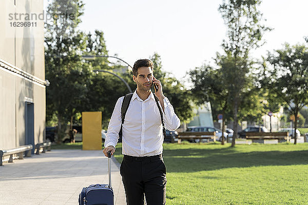 Geschäftsmann mit Koffer  der über sein Smartphone spricht  während er auf dem Fußweg geht