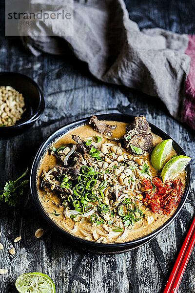 Schale mit roter Currysuppe mit Reisnudeln  Rindfleisch  Gemüse  Frühlingszwiebeln  Erdnüssen und Limette