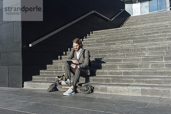 Geschäftsmann  der ein digitales Tablet benutzt  während er mit einem Elektroroller auf einer Treppe im Finanzbezirk sitzt